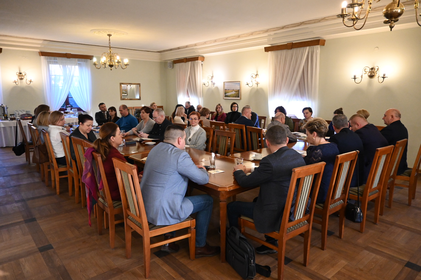 Na zdjęciu widoczna jest grupa osób siedzących przy dużym stole w sali konferencyjnej. Uczestnicy skupieni są na rozmowie. W tle znajdują się okna z zasłonami oraz bufet kawowy.