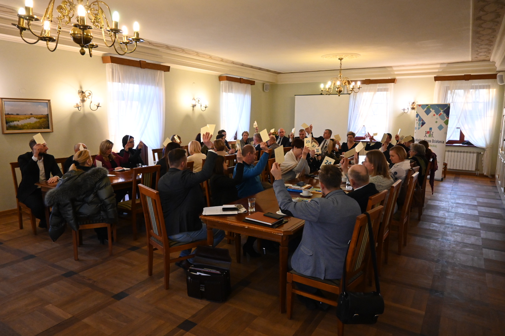 Na zdjęciu znajduje się grupa osób, siedzących przy stole. Podnoszą mandaty do głosowania. W tle znajduje się baner Lokalnej Grupy Działania Ziemi Siedleckiej.